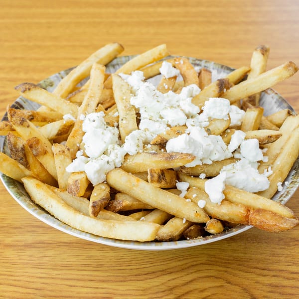 A bowl of golden french fries topped with crumbled feta cheese and herbs on a wooden table.