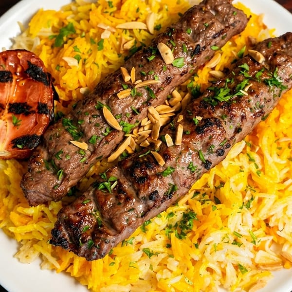 Two grilled meat skewers with herbs and almonds, served on yellow rice with a grilled tomato.
