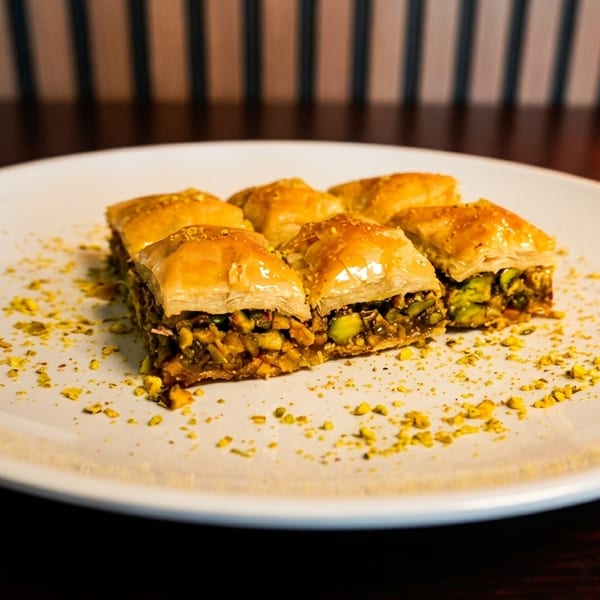 Six pieces of golden-brown baklava with pistachio filling, sprinkled with crushed pistachios on a white plate.