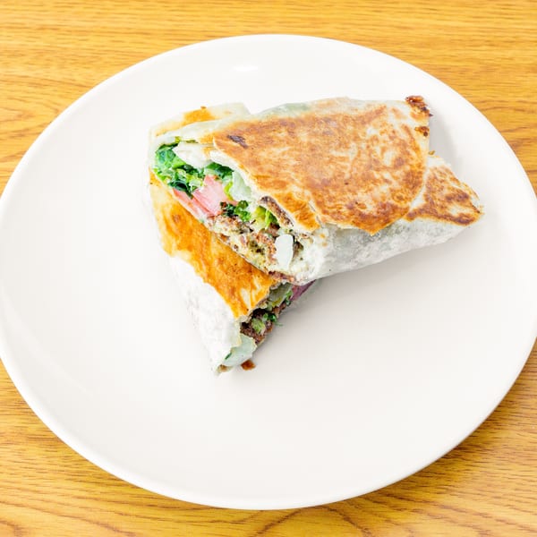A grilled wrap, cut in half, filled with meat, lettuce, and tomatoes, on a white plate.