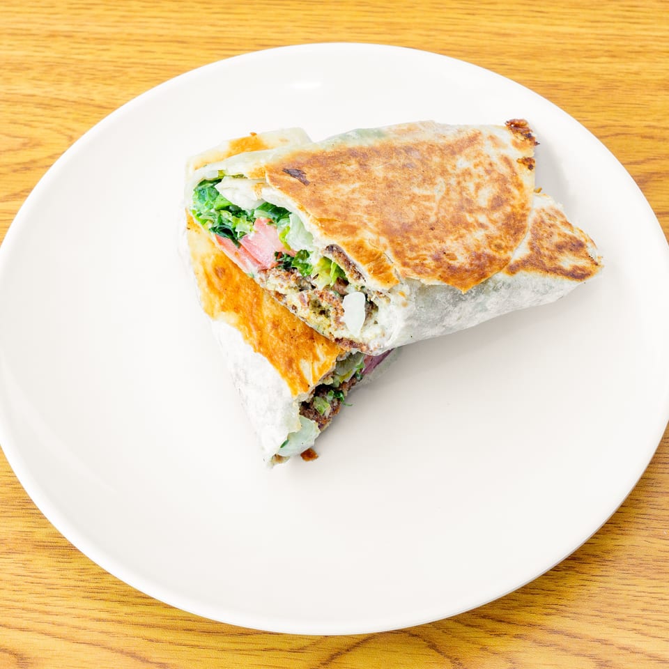 A grilled wrap, cut in half, filled with meat, lettuce, and tomatoes, on a white plate.