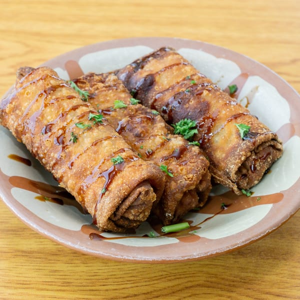 Three crispy, golden-brown egg rolls drizzled with dark sauce and garnished with parsley on a plate.