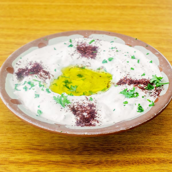 A bowl of creamy labneh drizzled with olive oil, sumac, and fresh parsley on a wooden table.
