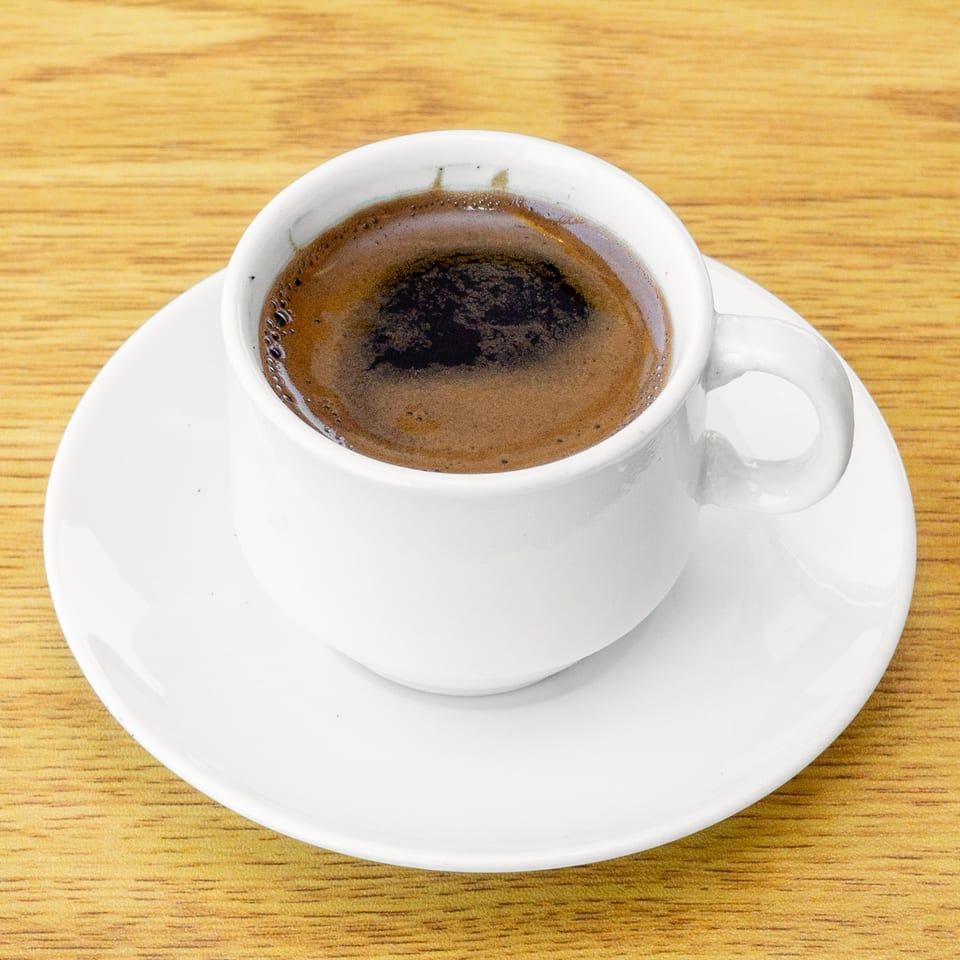 A white cup of dark Turkish coffee with foam on a matching saucer, on a wooden table.