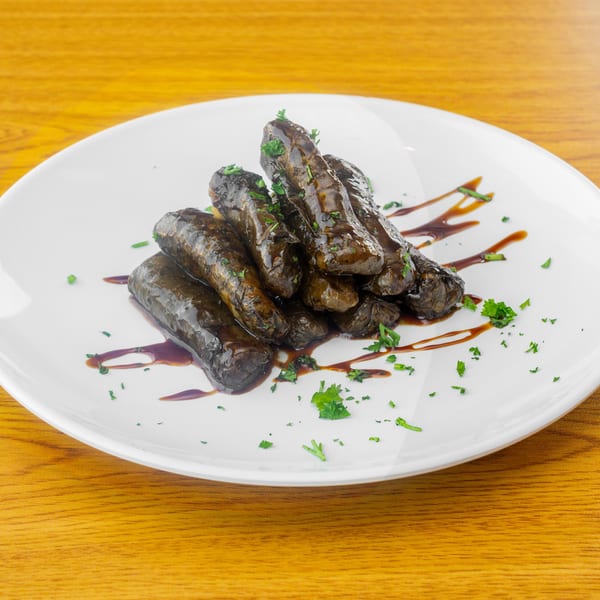 A white plate holds several dark, glossy dolmades drizzled with sauce and garnished with parsley.