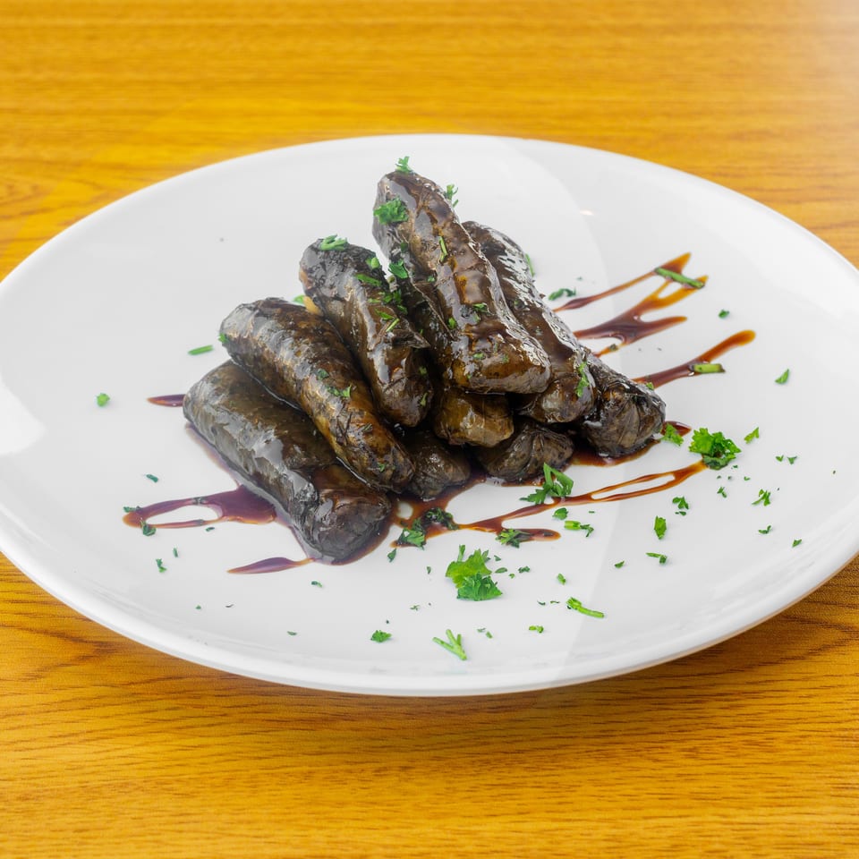 A white plate holds several dark, glossy dolmades drizzled with sauce and garnished with parsley.